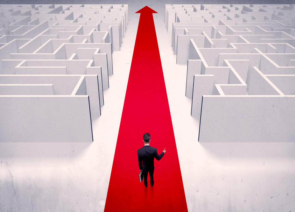 An adult elegant businessman standing on a red carpet arrow pointing ahead through a street with maze on two sides concept